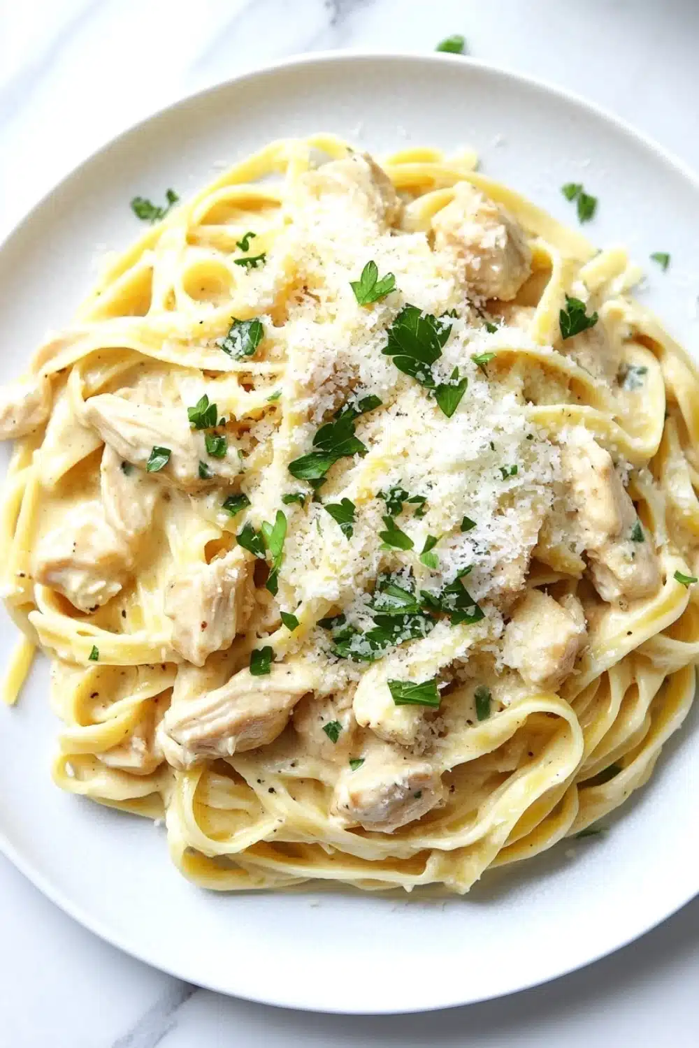 One pot chicken fettuccine - the image shows a plate of fettuccine alfredo pasta with chunks of chicken and parmesan cheese on top. the pasta is cooked al dente and is covered in a creamy white sauce. the dish is garnished with fresh parsley leaves. the plate is white and is sitting on a white marble countertop. the overall presentation of the dish is elegant and appetizing.
