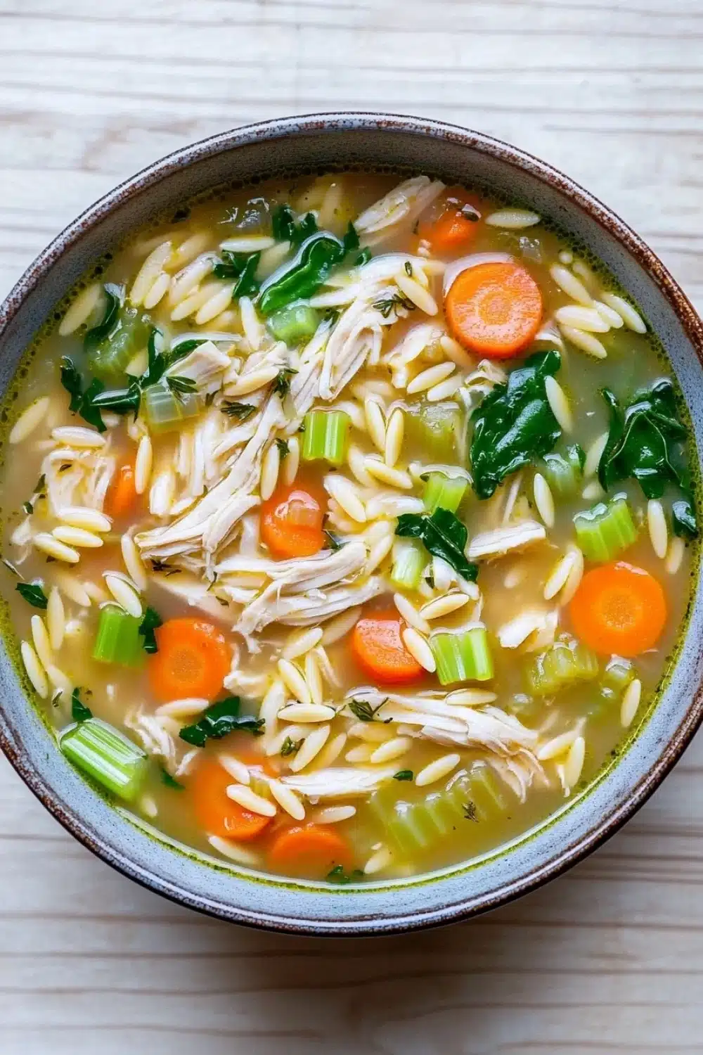 One pot chicken orzo soup - the image is a close-up of a bowl of soup. the soup appears to be a creamy yellow color with chunks of chicken and vegetables mixed in. the bowl is made of ceramic and has a brown rim. the broth is thick and creamy, and there are small pieces of green herbs scattered throughout the soup. there are also chunks of orange carrots and celery visible in the broth. the vegetables are bright green and appear to be fresh and healthy. the background is a light beige color, and the bowl is sitting on a white wooden surface.