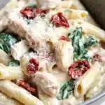 One pot creamy italian - the image is a close-up of a bowl of pasta dish. the pasta appears to be penne, which is a type of penne pasta. it is cooked al dente and is topped with chunks of chicken, spinach, and sun-dried tomatoes. the dish is garnished with grated parmesan cheese. the bowl is made of a light-colored ceramic material and is sitting on a wooden table. the background is blurred, so the focus is on the pasta and the dish.
