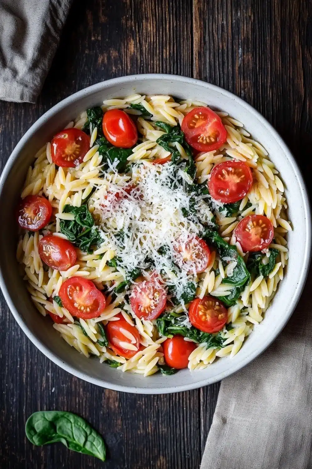 One pot pasta orzo - the image is a top view of a bowl of pasta salad. the bowl is made of white ceramic and is placed on a wooden table. the pasta is fusilli, which is a type of spiral-shaped pasta, and it is mixed with cherry tomatoes, spinach, and grated parmesan cheese. the tomatoes are bright red and appear to be ripe and juicy. the spinach is a vibrant green color and is scattered throughout the dish. there is a sprig of fresh basil on the side of the bowl, adding a pop of color to the image. a beige cloth napkin is visible in the background.
