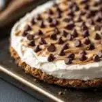 Peanut butter cookie crust dessert - the image shows a close-up of a dessert on a baking tray. the dessert appears to be a graham cracker crust with a layer of peanut butter frosting on top. the frosting is drizzled with chocolate chips, giving the dessert a crunchy texture. the tray is lined with parchment paper and there is a white napkin on the side. the background is a dark wooden table.