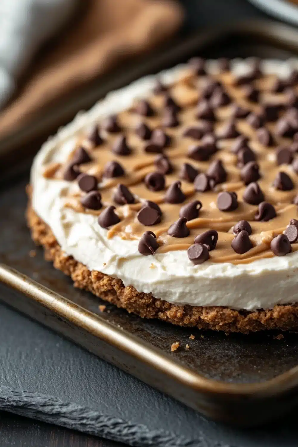 Peanut butter cookie crust dessert - the image shows a close-up of a dessert on a baking tray. the dessert appears to be a graham cracker crust with a layer of peanut butter frosting on top. the frosting is drizzled with chocolate chips, giving the dessert a crunchy texture. the tray is lined with parchment paper and there is a white napkin on the side. the background is a dark wooden table.