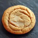 Peanut butter cookie no butter - the image is a close-up of a round cookie on a black slate surface. the cookie appears to be freshly baked and has a golden-brown color. it has a criss-cross pattern on the top, with small squares scattered throughout. the edges of the cookie are slightly curled, giving it a textured appearance. the background is blurred, making the cookie the focal point of the image.