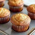 Plain yogurt muffin - the image shows a muffin tray with six freshly baked muffins in it. the muffins are golden brown in color and have a crumbly texture. they are arranged in a single layer in the tray, with the top layer slightly overlapping the bottom layer. the tray is lined with parchment paper and is placed on a white marble countertop. the background is blurred, but it appears to be a kitchen countertop with more muffins scattered around.