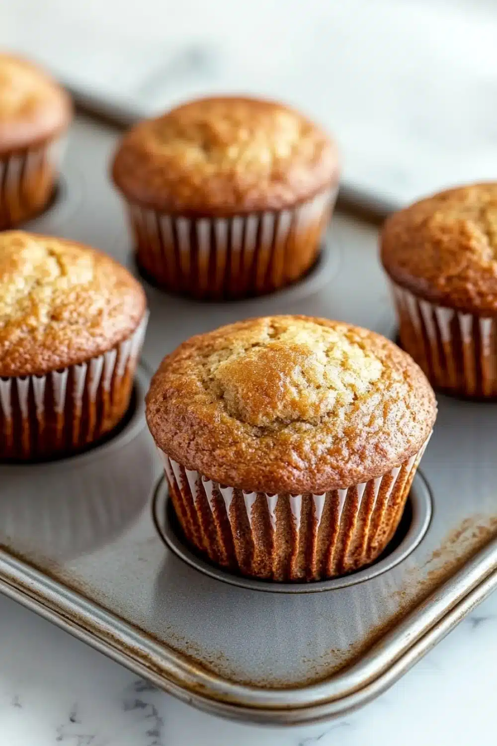 Plain yogurt muffin - the image shows a muffin tray with six freshly baked muffins in it. the muffins are golden brown in color and have a crumbly texture. they are arranged in a single layer in the tray, with the top layer slightly overlapping the bottom layer. the tray is lined with parchment paper and is placed on a white marble countertop. the background is blurred, but it appears to be a kitchen countertop with more muffins scattered around.