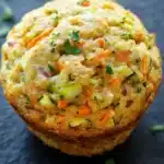 Protein veggie muffin - the image is a close-up of a muffin on a black surface. the muffin appears to be freshly baked and has a golden-brown crust. it is filled with a mixture of vegetables, including carrots, zucchini, and onions, and is garnished with chopped parsley. the vegetables are arranged in a circular pattern on top of the muffin, with some overlapping each other. the background is blurred, but it seems to be a dark, textured surface.