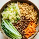 Ramen noodle skillet ground beef - the image is a close-up of a bowl of noodles and vegetables. the bowl is made of a dark brown ceramic material and is placed on a wooden table. the noodles are yellow and appear to be ramen-like. on top of the noodles, there is a pile of ground beef, which is brown and appears to be seasoned with herbs and spices. next to the beef, there are sliced green onions and sliced carrots. the vegetables are arranged in a colorful and appetizing manner. the overall color scheme of the dish is warm and inviting.