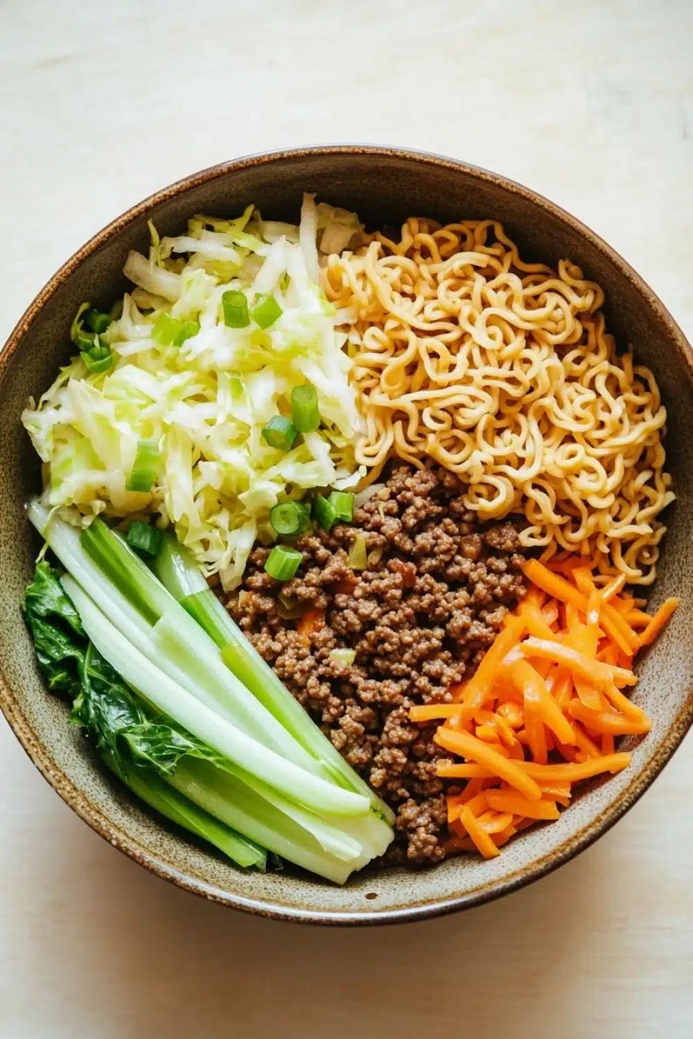 Ramen noodle skillet ground beef - the image is a close-up of a bowl of noodles and vegetables. the bowl is made of a dark brown ceramic material and is placed on a wooden table. the noodles are yellow and appear to be ramen-like. on top of the noodles, there is a pile of ground beef, which is brown and appears to be seasoned with herbs and spices. next to the beef, there are sliced green onions and sliced carrots. the vegetables are arranged in a colorful and appetizing manner. the overall color scheme of the dish is warm and inviting.