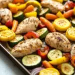 Sheet pan chicken and veggies - the image shows a baking tray filled with grilled chicken and vegetables. the chicken is cooked to a golden brown color and appears to be seasoned with herbs and spices. the vegetables are cut into small pieces and are arranged in a circular pattern on the tray. there are also sliced zucchini, red and yellow bell peppers, and red onions scattered throughout the tray, adding a pop of color to the dish. the tray is sitting on a white countertop.