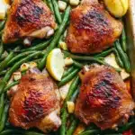Sheet pan chicken green beans - the image shows a rectangular baking dish filled with grilled chicken and green beans. the chicken is golden brown and appears to be seasoned with herbs and spices. the green beans are bright green and look fresh and vibrant. there are two lemon wedges on the side of the dish, adding a pop of color to the dish. the dish is sitting on a wooden table, and the background is blurred, making the dish the focal point of the image.