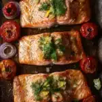 Sheet pan lemon brown butter salmon - the image shows two pieces of salmon fillets on a baking tray. the salmon is cooked to a golden brown color and is garnished with fresh dill and lemon zest. there are also sliced red onions and cherry tomatoes scattered around the tray, adding a pop of color to the dish. the tray is lined with parchment paper and there are a few sprigs of dill scattered around it. the overall presentation of the dish is appetizing and visually appealing.