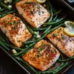 Sheet pan salmon green beans - the image shows a baking tray with four pieces of salmon and green beans. the salmon is cooked to a golden brown color and appears to be seasoned with herbs and spices. the green beans are long and slender, and they are arranged in a single layer on the tray. there are two lemon wedges on either side of the salmon, and a few garlic cloves scattered around the tray, adding a pop of color to the dish. the tray is placed on a wooden table, and there is a white plate in the background.