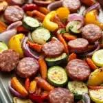 Sheet pan smoked sausage and veggies - the image shows a tray of roasted vegetables on a baking sheet. the vegetables include sliced zucchini, red onions, yellow bell peppers, and red onions. the sausage is cooked to a golden brown color and appears to be seasoned with herbs and spices. the tray is lined with parchment paper and is sitting on a wooden table. the background is blurred, but it seems to be a kitchen countertop.