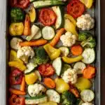 Sheet pan vegetables air fryer - the image shows a rectangular baking tray filled with roasted vegetables. the vegetables are arranged in a single layer on the tray, with some overlapping each other. the tray is lined with parchment paper and is placed on a wooden table. the vegetables include zucchini, bell peppers, broccoli, cauliflower, and cherry tomatoes. the colors of the vegetables are vibrant and the dish looks fresh and appetizing. the background is blurred, but it appears to be a kitchen countertop.