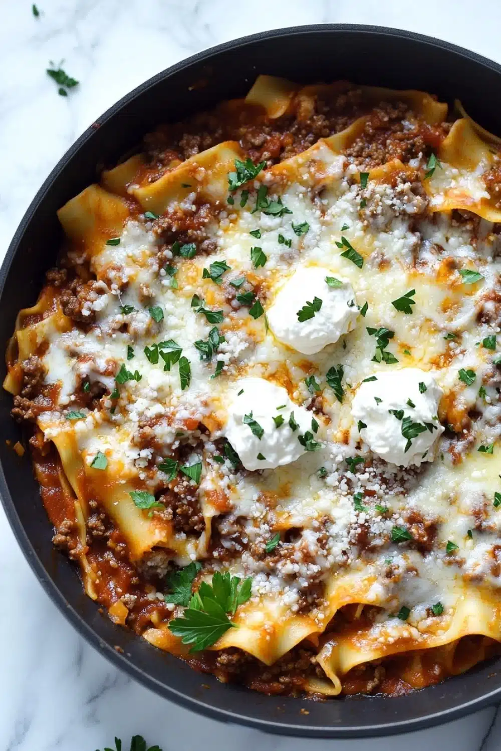 Skillet beef lasagna - the image shows a close-up of a dish in a black cast iron skillet. the dish appears to be a lasagna or lasagna roll-up, with a layer of ground beef and tomato sauce on top. the top of the dish is covered in melted cheese and sprinkled with chopped parsley. the skillet is sitting on a white marble countertop.