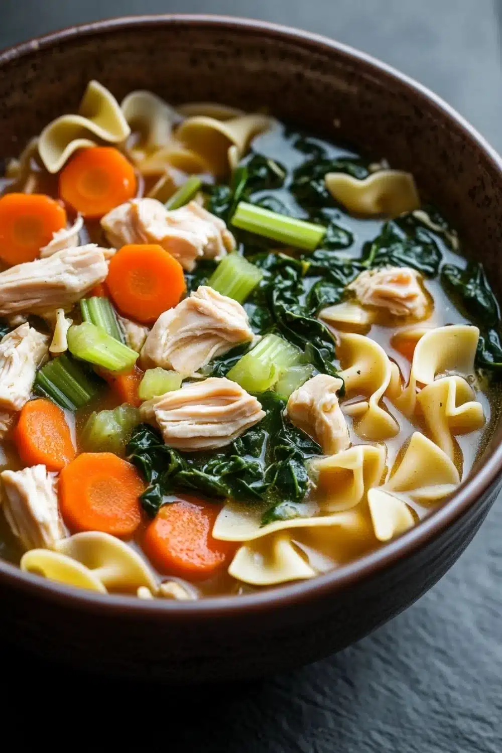 Skillet chicken noodle soup - the image is a close-up of a bowl of soup. the soup appears to be a type of noodle soup with chunks of chicken, carrots, and greens mixed in. the noodles are long and thin, and the vegetables are bright orange and green. the broth is a light brown color and looks thick and creamy. the bowl is made of dark brown ceramic and is sitting on a dark grey countertop. the background is blurred, making the soup the focal point of the image.