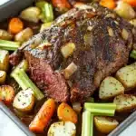 Slow cooker beef chuck roast - the image shows a large rectangular baking tray filled with a dish of roast beef and vegetables. the roast beef is cooked medium-rare and appears to be seasoned with herbs and spices. the vegetables include carrots, celery, and potatoes. the tray is sitting on a white marble countertop with a blue and white striped napkin on the side. the dish looks freshly cooked and ready to be served.