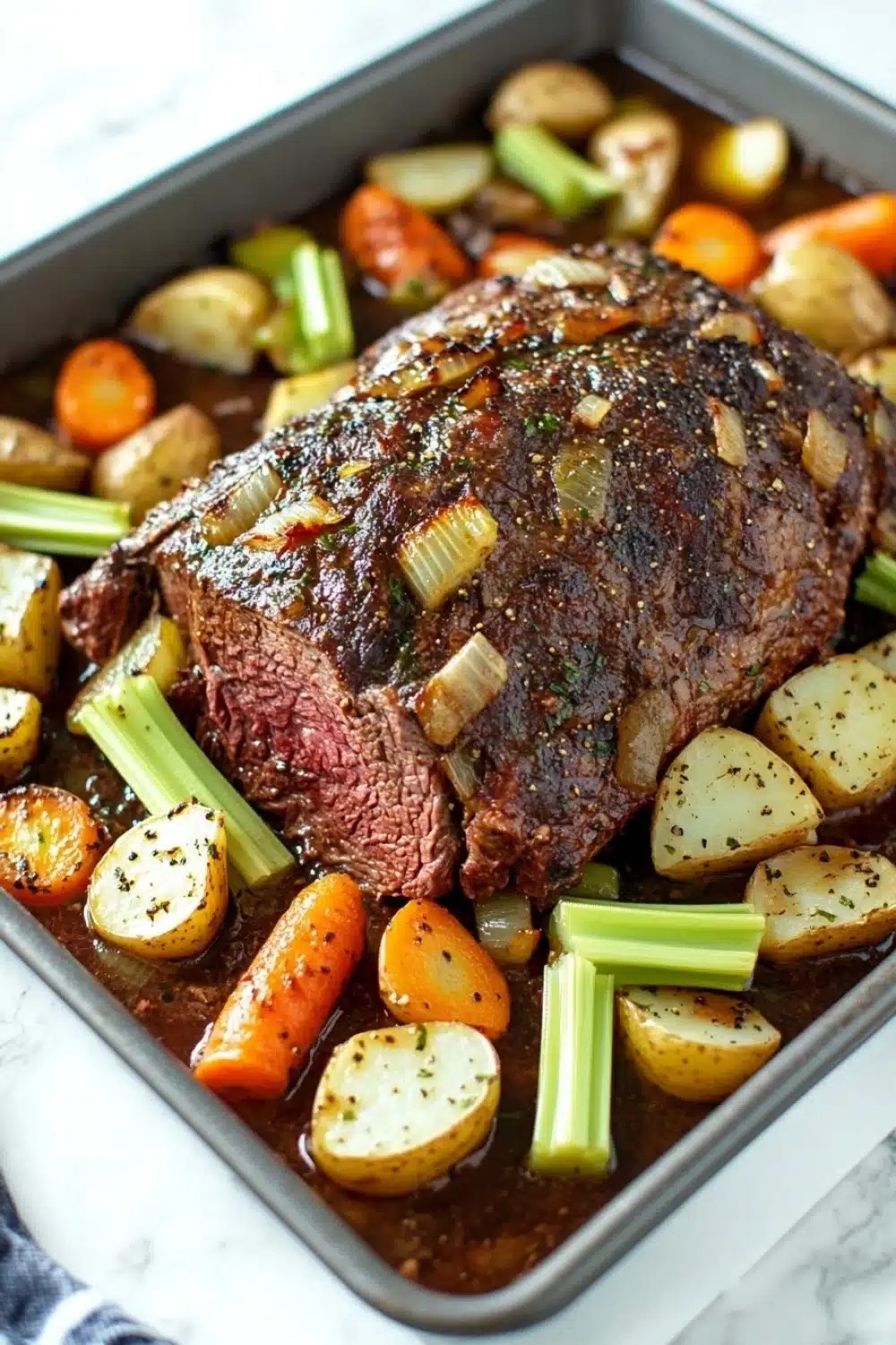 Slow cooker beef chuck roast - the image shows a large rectangular baking tray filled with a dish of roast beef and vegetables. the roast beef is cooked medium-rare and appears to be seasoned with herbs and spices. the vegetables include carrots, celery, and potatoes. the tray is sitting on a white marble countertop with a blue and white striped napkin on the side. the dish looks freshly cooked and ready to be served.