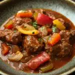 Slow cooker beef goulash - the image is a close-up of a dish in a bowl. the dish appears to be a beef stew with chunks of beef, red and yellow bell peppers, onions, and herbs. the beef is cooked in a tomato-based sauce and is garnished with chopped parsley. the bowl is sitting on a dark wooden table. the overall color palette of the dish is warm and inviting.