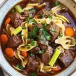 Slow cooker beef noodle - the image is a close-up of a bowl of beef noodle soup. the soup is a rich brown color and appears to be thick and hearty. it is filled with chunks of beef, carrots, celery, and noodles. the beef is cooked to a medium-rare and is garnished with a sprig of fresh parsley. the noodles are thin and fluffy, and the soup is thick and thick. the bowl is sitting on a white marble countertop.