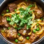 Slow cooker beef noodle curry - the image is a close-up of a bowl of beef stew. the stew is made with chunks of beef, onions, and peas, and is garnished with a sprig of fresh cilantro. the beef appears to be cooked medium-rare and has a reddish-brown color. the onions are thin and yellow, and the peas are bright green. the dish is served in a dark brown bowl with a dark rim. the background is a dark grey countertop.