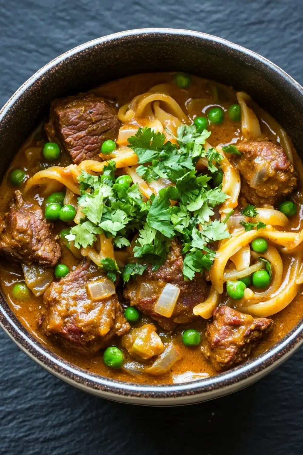 Slow cooker beef noodle curry - the image is a close-up of a bowl of beef stew. the stew is made with chunks of beef, onions, and peas, and is garnished with a sprig of fresh cilantro. the beef appears to be cooked medium-rare and has a reddish-brown color. the onions are thin and yellow, and the peas are bright green. the dish is served in a dark brown bowl with a dark rim. the background is a dark grey countertop.