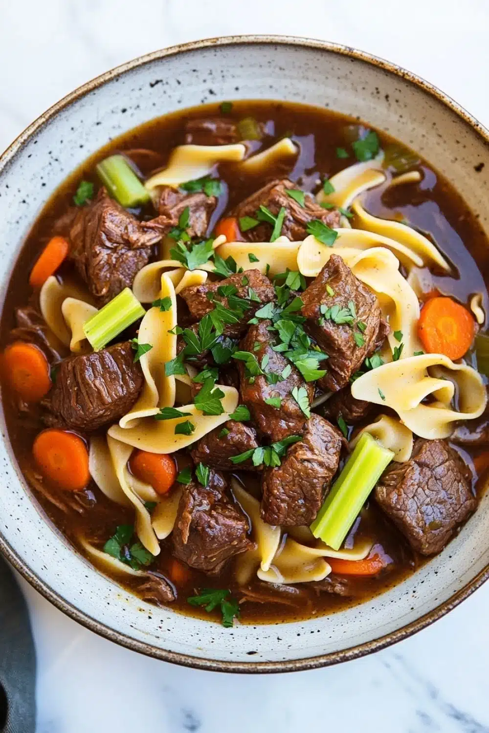 Slow cooker beef noodle - the image is a close-up of a bowl of beef noodle soup. the soup is a rich brown color and appears to be thick and hearty. it is filled with chunks of beef, carrots, celery, and noodles. the beef is cooked to a medium-rare and is garnished with a sprig of fresh parsley. the noodles are thin and fluffy, and the soup is thick and thick. the bowl is sitting on a white marble countertop.
