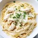 Slow cooker chicken alfredo - the image is a close-up of a bowl of pasta dish. the pasta appears to be fettuccine alfredo, with a creamy white sauce and chunks of chicken mixed in. the dish is garnished with chopped parsley and grated parmesan cheese. the bowl is white and is sitting on a white marble countertop. there is a fork resting on the side of the bowl.