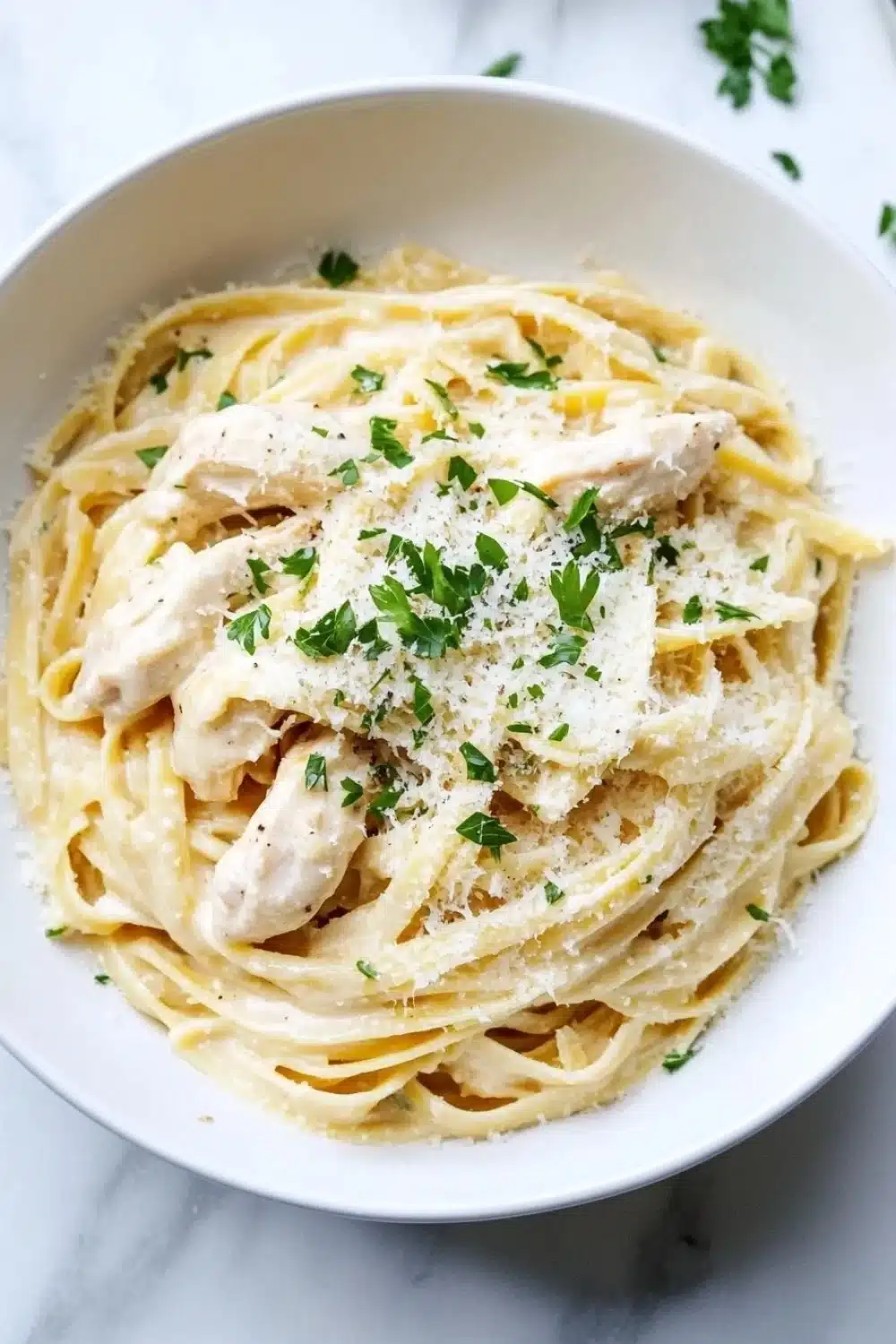 Slow cooker chicken alfredo - the image is a close-up of a bowl of pasta dish. the pasta appears to be fettuccine alfredo, with a creamy white sauce and chunks of chicken mixed in. the dish is garnished with chopped parsley and grated parmesan cheese. the bowl is white and is sitting on a white marble countertop. there is a fork resting on the side of the bowl.