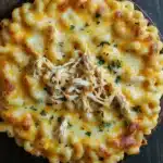 Slow cooker chicken mac and cheese - the image is a close-up of a dish of macaroni and cheese. the dish is in a black bowl and is placed on a wooden table. the top of the dish is covered in melted cheese and there are chunks of shredded chicken scattered throughout. the cheese is a golden yellow color and appears to be melted and bubbly. there are also some chopped parsley scattered around the dish, adding a pop of green color to the dish.