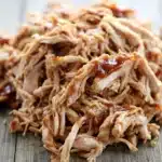 Slow cooker chicken pulled - the image shows a pile of shredded chicken on a wooden surface. the chicken appears to be cooked and has a golden brown color. it is covered in a thick layer of bbq sauce, which is a reddish-brown color and is drizzled over the top of the chicken. the shredded chicken is arranged in a pile, with some overlapping each other. the background is blurred, but it seems to be a wooden table or countertop.