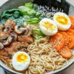 Slow cooker chicken ramen - the image is a close-up of a bowl of ramen. the bowl is made of ceramic and is sitting on a wooden table. the ramen is a light brown color and appears to be thick and fluffy. on top of the ramen, there are sliced mushrooms, sliced carrots, and a hard-boiled egg. there are also some sesame seeds sprinkled on top. the dish is garnished with green onions and sesame seed. the background is blurred, but it seems to be a kitchen countertop.