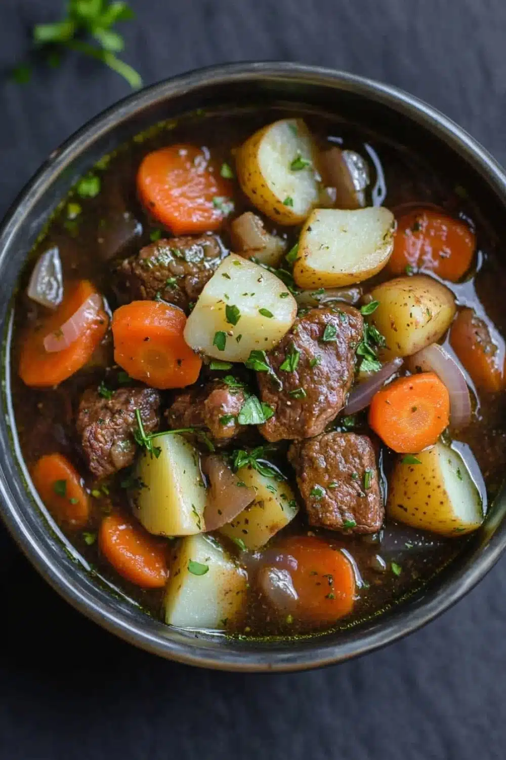 Slow cooker poor mans beef stew - the image is a close-up of a bowl of beef stew. the stew is made with chunks of beef, potatoes, carrots, onions, and herbs. the beef is cooked medium-rare and appears to be seasoned with herbs and spices. the potatoes are cut into small cubes and are scattered throughout the stew, with some of them overlapping each other. the carrots are bright orange and appear to be ripe and juicy. the onions are sliced and are also diced. the herbs are finely chopped and add a pop of green color to the dish. the bowl is made of stainless steel and is sitting on a dark grey surface.