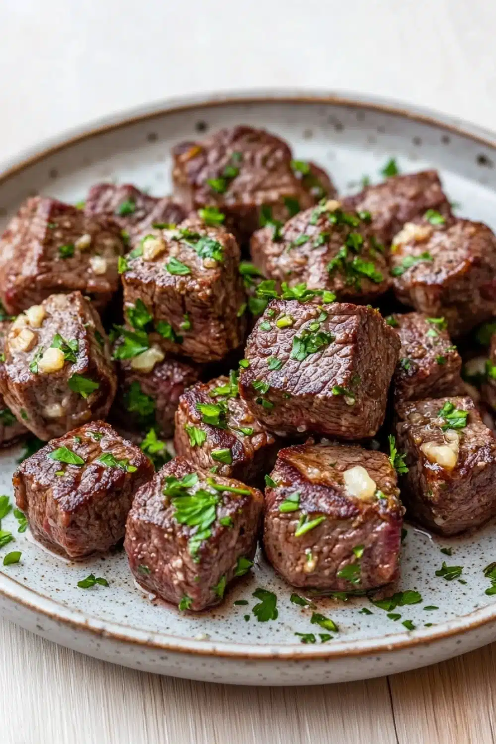 Steak bites with garlic butter crockpot - the image shows a plate of cooked beef cubes. the cubes are dark brown in color and appear to be seasoned with herbs and spices. they are arranged in a circular pattern on the plate, with some overlapping each other. the plate is white with a light blue rim and is placed on a wooden table. the background is blurred, but it appears to be a kitchen countertop.
