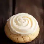 Sugar cookie and frosting - the image is a close-up of a round cookie with a swirl of white frosting on top. the cookie appears to be freshly baked and is placed on a wooden surface. the frosting is swirled in a spiral pattern and is spread evenly across the top of the cookie. the background is dark and out of focus, making the cookie the focal point of the image.