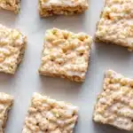 Sugar cookie rice crispy - the image shows a group of square-shaped rice krispy treats arranged on a white marble surface. the treats are golden brown in color and have a crumbly texture. they are topped with a generous layer of white cream cheese frosting, which is drizzled over the top of each treat. the frosting appears to be a light yellow color and has small bits of nuts scattered throughout. the background is blurred, making the treats the focal point of the image.