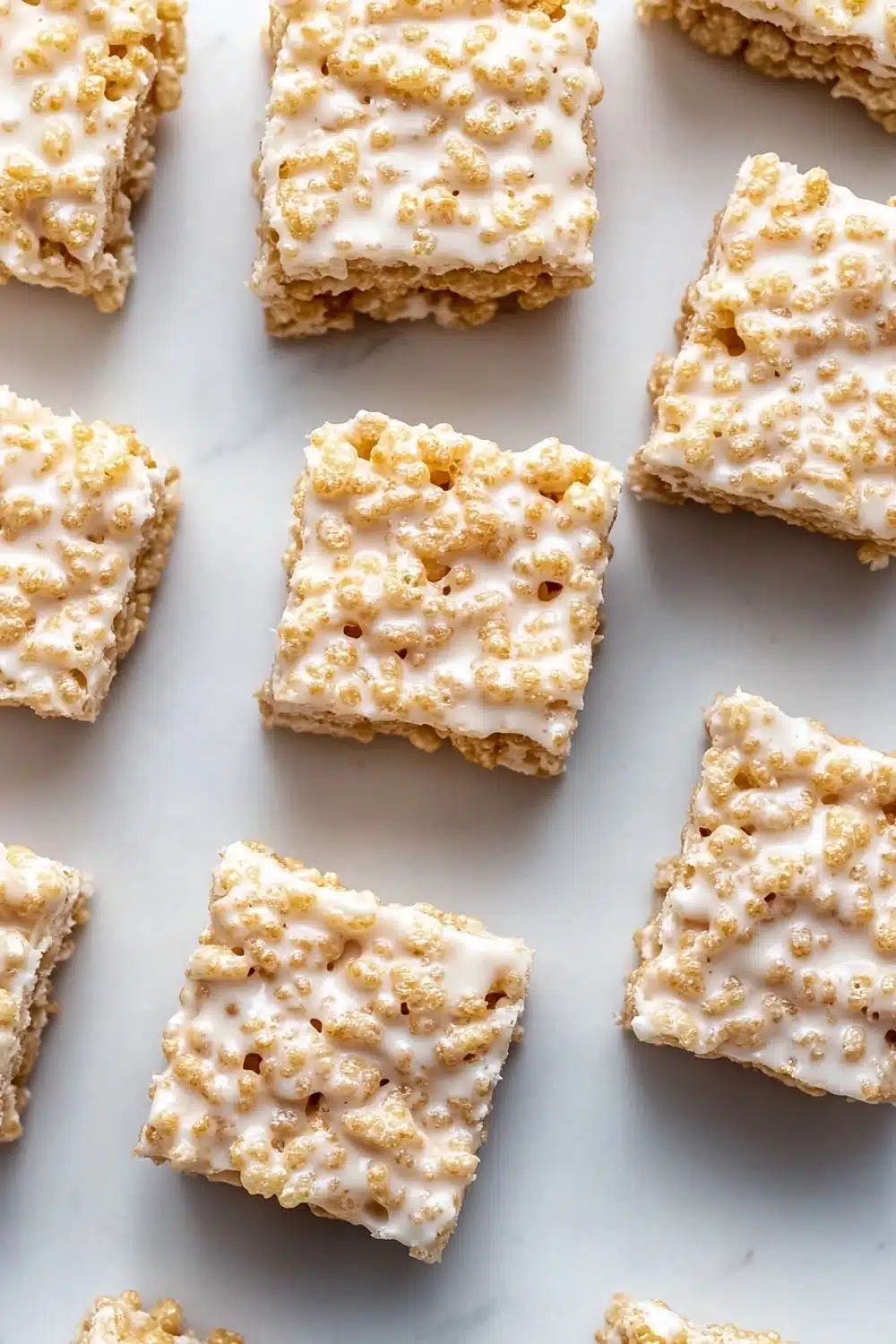 Sugar cookie rice crispy - the image shows a group of square-shaped rice krispy treats arranged on a white marble surface. the treats are golden brown in color and have a crumbly texture. they are topped with a generous layer of white cream cheese frosting, which is drizzled over the top of each treat. the frosting appears to be a light yellow color and has small bits of nuts scattered throughout. the background is blurred, making the treats the focal point of the image.