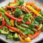 Thai vegetable air fryer - the image is a close-up of a plate of stir-fry dish. the dish is made up of various vegetables, including bell peppers, broccoli, and snow peas. the vegetables are cut into thin strips and are arranged in a colorful and appetizing manner. the plate is white with a speckled pattern and is garnished with sesame seeds and fresh cilantro. the background is dark, making the colors of the vegetables stand out.