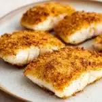 Tilapia in air fryer - the image shows a plate of four pieces of fish fillets on a white plate. the fillets are golden brown and appear to be seasoned with a crumbly breadcrumb topping. the plate is round and has a light brown rim. the background is blurred, but it appears to be a kitchen countertop.
