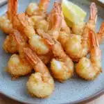 Trader joes shrimp tempura air fryer - the image shows a plate of cooked shrimp on a wooden table. the shrimp are golden brown and appear to be crispy on the outside. they are arranged in a circular pattern on the plate, with some overlapping each other. there is a slice of lemon on the side of the plate. the plate is a light brown color with a speckled texture. the background is blurred, but it appears to be a kitchen countertop.