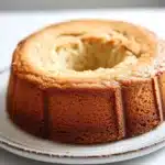 Vanilla cake no icing - the image is of a round cake on a white cake stand. the cake appears to be freshly baked and has a golden brown color. the top of the cake is covered in a light dusting of powdered sugar, giving it a shiny appearance. the stand is made of metal and is placed on a marble countertop. the background is blurred, making the cake the focal point of the image.