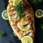 Whole tilapia in the air fryer - the image shows a whole fish on a black slate board. the fish appears to be a tilapia, with a golden-brown color and a crispy texture. it is garnished with sliced lemons and a sprig of fresh parsley. there are also a few whole lemons scattered around the fish, adding a pop of color to the dish. the background is dark, making the fish stand out.