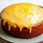Yellow lemon cake design - the image is of a round cake on a white plate. the cake appears to be a chocolate cake with a golden brown crust. on top of the cake, there is a drizzle of yellow icing that is dripping down the sides. the icing is drizzled over the top, creating a smooth and glossy texture. the plate has a speckled pattern around the edges. the background is a plain white surface.