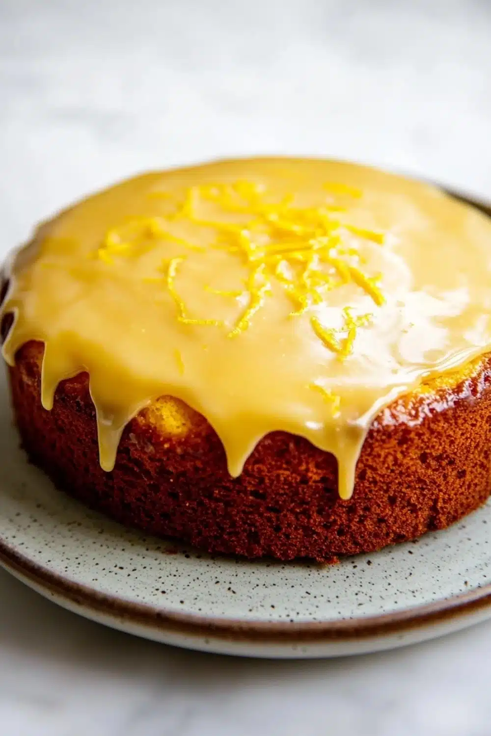 Yellow lemon cake design - the image is of a round cake on a white plate. the cake appears to be a chocolate cake with a golden brown crust. on top of the cake, there is a drizzle of yellow icing that is dripping down the sides. the icing is drizzled over the top, creating a smooth and glossy texture. the plate has a speckled pattern around the edges. the background is a plain white surface.