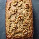 Zucchini bread almond flour - the image is a close-up of a freshly baked loaf of bread on a black slate surface. the bread appears to be golden brown in color and has a crumbly texture. on top of the bread, there are several walnuts scattered across the surface, giving it a crunchy appearance. the walnuts are arranged in a circular pattern, with some overlapping each other. the loaf is slightly tilted to the side, and the edges are slightly curled. the background is blurred, making the bread the focal point of the image.
