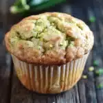 Zucchini lemon muffin - the image is of a freshly baked zucchini muffin on a wooden table. the muffin is golden brown in color and has a crumbly texture. it is topped with a layer of zucchinis and sprinkled with chopped herbs. in the background, there is a whole zucchi and a few scattered herbs, adding a pop of green color to the image. the overall mood of the image is rustic and appetizing.