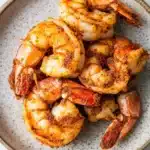 Air fryer carribean jerk shrimp - the image shows a plate of cooked shrimp on a white marble countertop. the shrimp are arranged in a circular pattern on the plate, with some overlapping each other. they are covered in a dark brown sauce, which appears to be seasoned with spices and herbs. the sauce is drizzled over the shrimp, giving them a glossy sheen. the plate is round and has a speckled texture. the overall appearance of the dish is appetizing and ready to eat.
