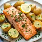 Air fryer salmon and potatoes - the image shows a plate of food with a piece of salmon on it. the salmon is cooked to a golden brown color and is garnished with fresh herbs and lemon wedges. the plate is white and round, and there are small potatoes scattered around the plate. the background is a white marble countertop with a gray cloth napkin on the side. the overall presentation of the dish is elegant and appetizing.