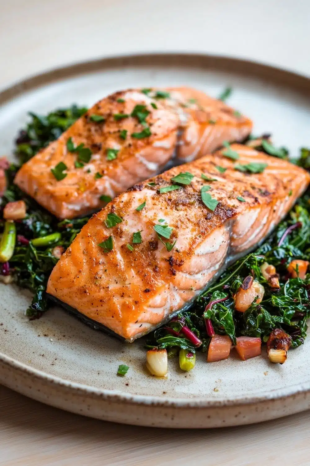 Air fryer salmon and swiss chard - the image shows two pieces of salmon on a plate. the salmon is cooked to a golden brown color and is garnished with fresh herbs. the plate is made of light-colored wood and has a textured surface. on top of the salmon, there is a bed of green leafy vegetables, including kale, red onions, and chickpeas. the vegetables are arranged in a circular pattern around the salmon. the background is blurred, but it appears to be a wooden table.