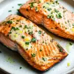 Air fryer salmon garlic butter - the image shows two pieces of salmon on a white plate. the salmon is cooked to a golden brown color and is garnished with chopped parsley and lemon wedges. the plate is sitting on a wooden table with a blurred background. the fish appears to be seasoned with herbs and spices, giving it a savory flavor.